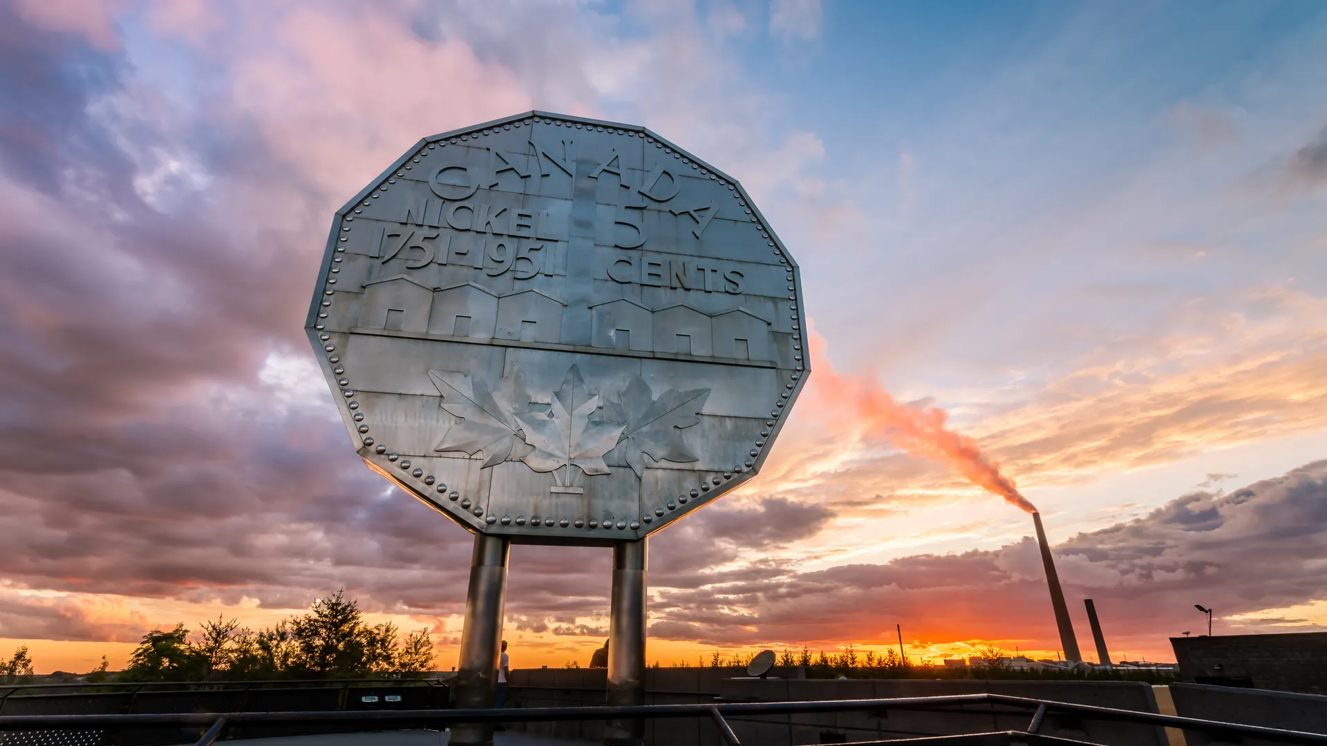 A photo of the Big Nickel in Sudbury, Ontario, Canada at sunset with a cloudy, multi-colored sky in the background and the famous Smoke Stack visible in the distance. A view that is uniquely Sudbury.